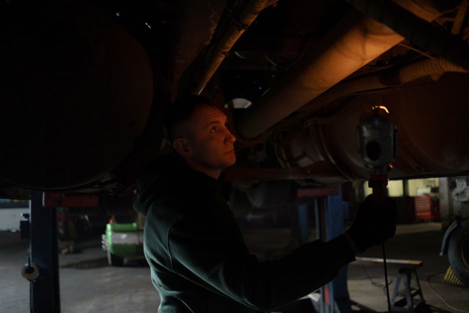 Mechanic working in dimly lit garage, inspecting car's undercarriage with a portable light source.