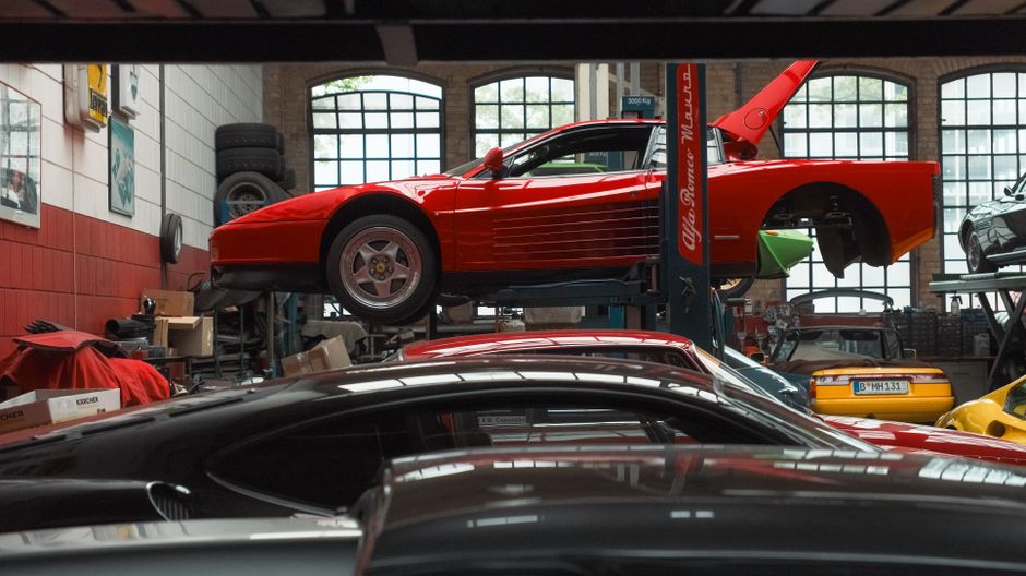 A vibrant red sports car is elevated on a lift inside an auto repair garage, surrounded by various vehicles and equipment.